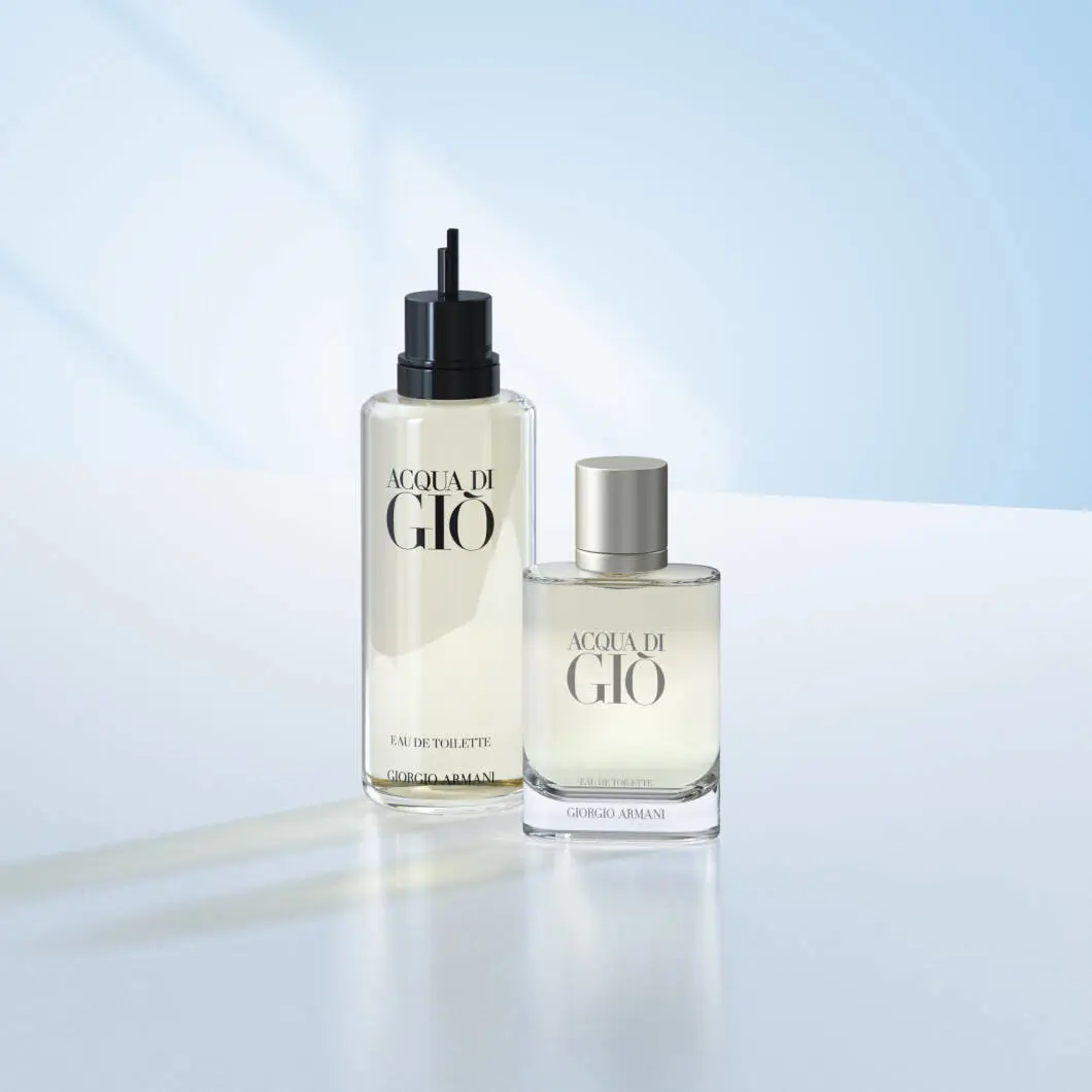 To Acqua di Giò-parfumeprodukter fra Giorgio Armani står på en lys overflade foran en blød lyseblå baggrund. Til venstre ses en høj refillflaske med sort pumpe, og til højre står den klassiske parfumeflaske med sølvfarvet låg. Billedet har et rent og elegant udtryk med fokus på luksusparfume og refill-løsninger.