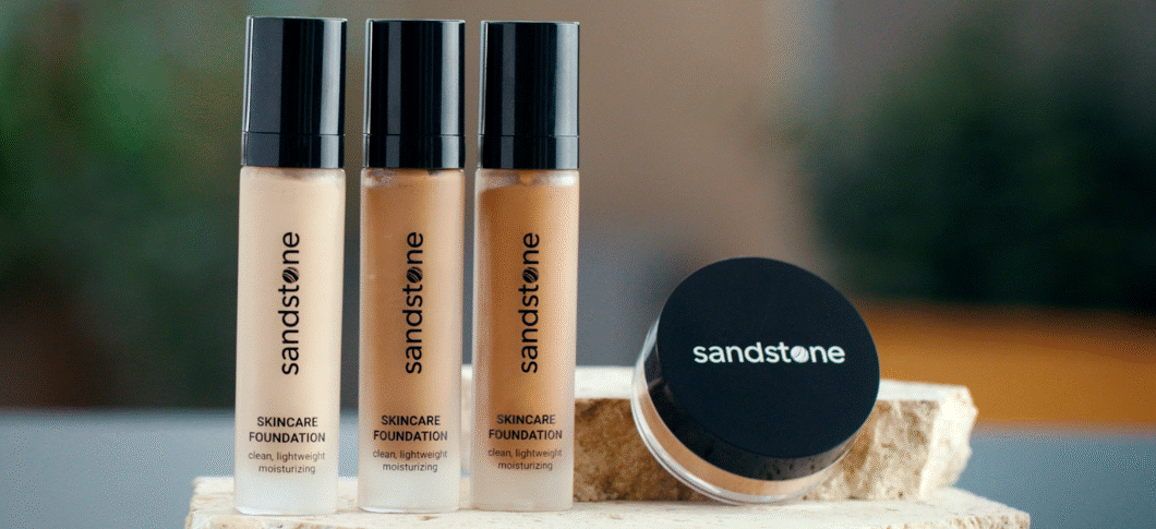 Her ses et billede med tre foundation-flasker og en rund beholder fra Sandstone placeret på en lys overflade. På flaskerne står teksten “sandstone Skincare Foundation”, og på den runde beholder står “sandstone”. Produkterne er i forskellige hudtoner med sorte låg.