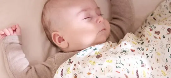 Sovende baby ligger under en lys dyne med små farverige motiver i rolige, beige omgivelser.