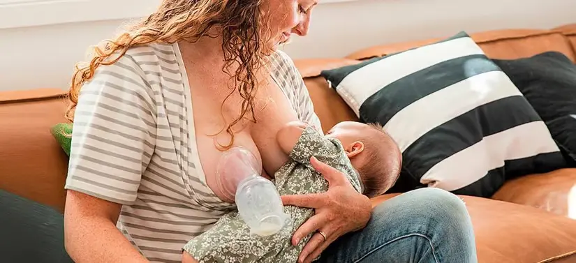 Her ses en kvinde som ammer på det ene bryst og har en brystpumpe på det andet. Hun sidder i en brun sofa.