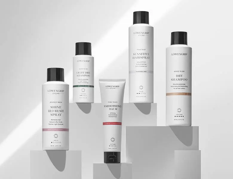 Her ses et billede med flere hårprodukter fra Løwengrip placeret på podier mod en lys baggrund. På produkterne står blandt andet “Shine Refresh Spray”, “Light Dry Shampoo”, “Sensitive Hairspray”, “Smoothing Balm” og “Dry Shampoo”. Produkterne er i hvide emballager med sort låg.