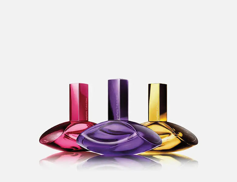 Her ses et billede med tre parfumeflakoner fra Calvin Klein placeret side om side på en lys, reflekterende overflade mod en lys baggrund. Flakonerne er i farverne pink, lilla og gul med blanke overflader og aflange låg.
