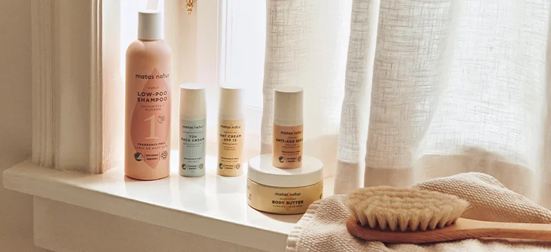 Her ses et stemningsfuldt billede med produkter fra Matas Natur placeret i en lys vindueskarm. Sortimentet omfatter blandt andet Low-Poo Shampoo, ansigtspleje, Day Cream SPF 15, Anti-Age SPF og Body Butter. Ved siden af produkterne ligger en kropsbørste og et tekstil, mens det varme dagslys og de naturlige materialer skaber et roligt og plejende udtryk.