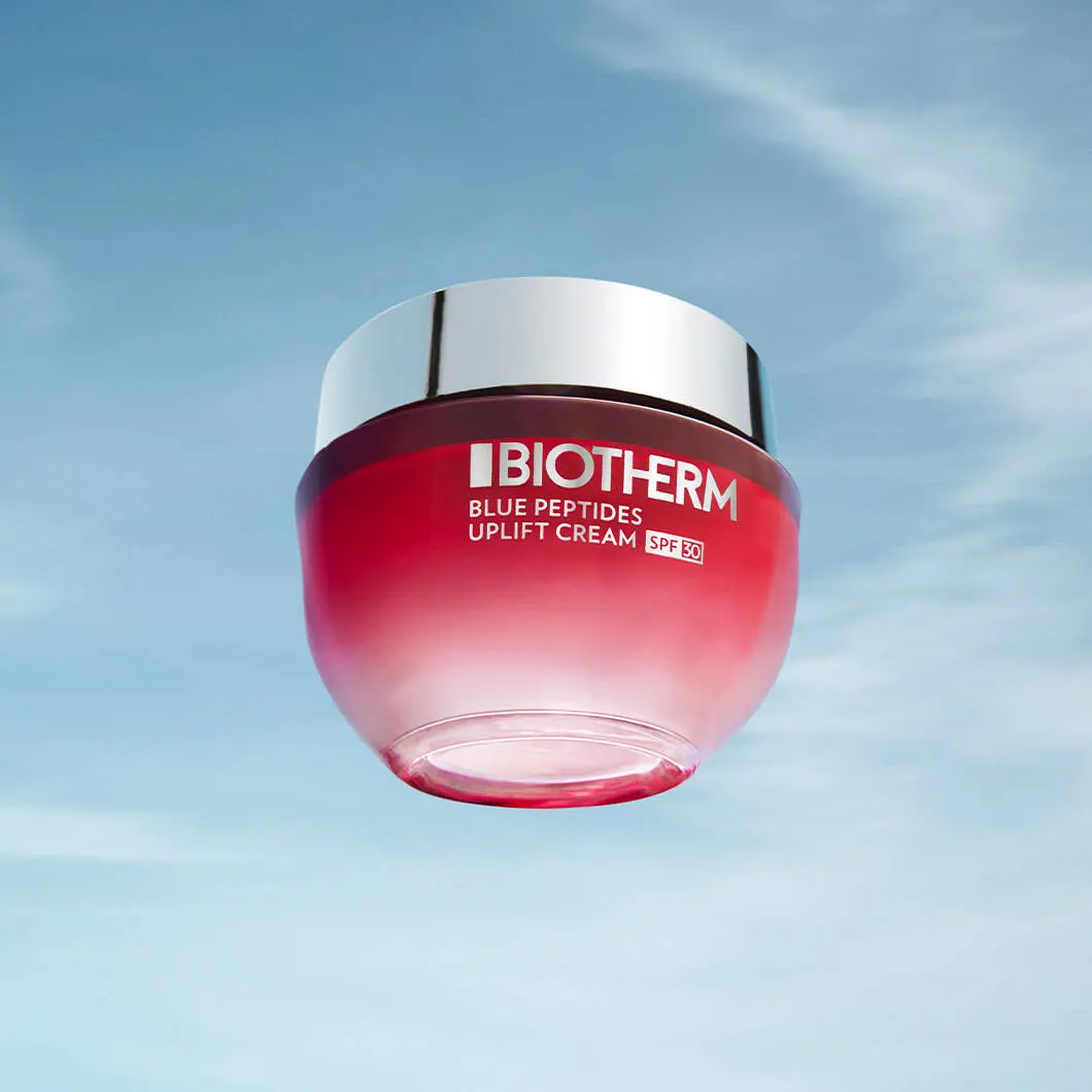 Et produktbillede af en Biotherm Blue Peptides Uplift Cream med SPF 30 placeret foran en lys blå himmel med lette skyer. Produktet svæver midt i billedet og består af en rund cremebeholder i røde og rosa nuancer med et blankt sølvfarvet låg. På forsiden står Biotherms logo samt teksten “Blue Peptides Uplift Cream SPF 30” med hvide bogstaver. Det bløde dagslys og den enkle baggrund giver billedet et friskt og moderne udtryk med fokus på hudpleje, opstramning og solbeskyttelse.