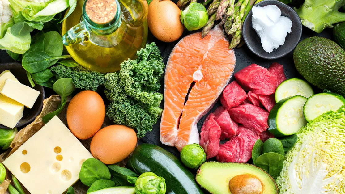 Her ses forskellige madvarer som laks, kød, æg, avocado, grøntsager og olie arrangeret på et bord.