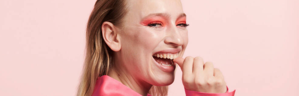 Her ses en lyserød baggrund med en glad kvindes ansigt set fra siden. Hun har en pink skjorte og øjenmakeup på. Hun har spidsen af sin tommelfinger i munden.