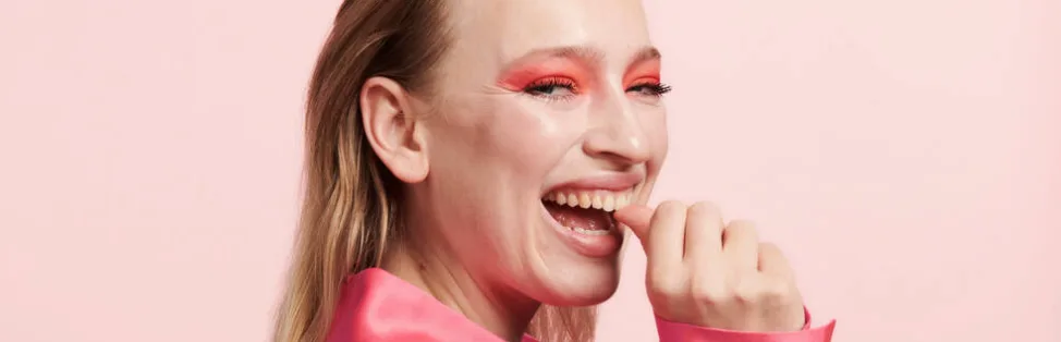 Her ses en lyserød baggrund med en glad kvindes ansigt set fra siden. Hun har en pink skjorte og øjenmakeup på. Hun har spidsen af sin tommelfinger i munden.
