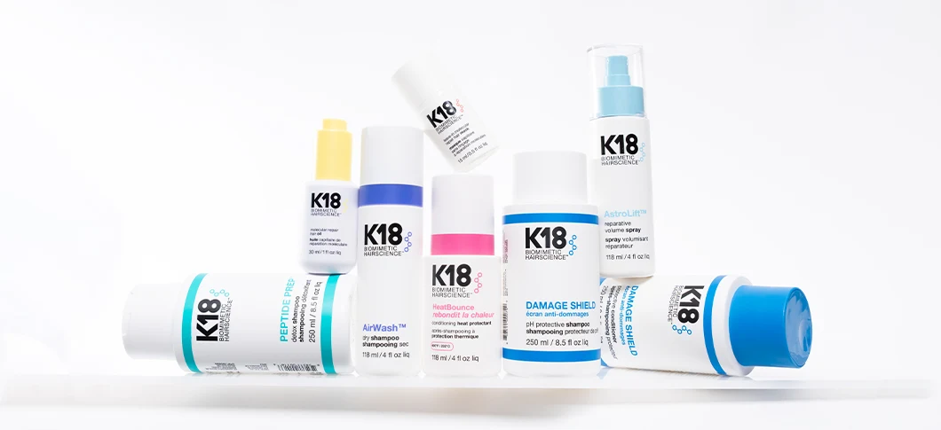 Her ses et udvalg af K18-hårplejeprodukter i hvide emballager med detaljer i blå, pink, gul og turkis. Sortimentet omfatter blandt andet AirWash Dry Shampoo, HeatBounce, Damage Shield, AstroLift Volume Spray og Peptide Prep. Produkterne er arrangeret samlet mod en lys baggrund.