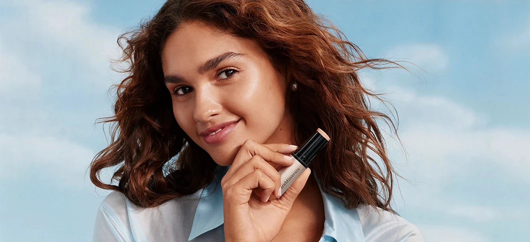 Her ses et billede af en person mod en himmelblå baggrund med skyer. Personen holder et makeup-produkt fra bareMinerals op ved ansigtet og smiler mod kameraet.