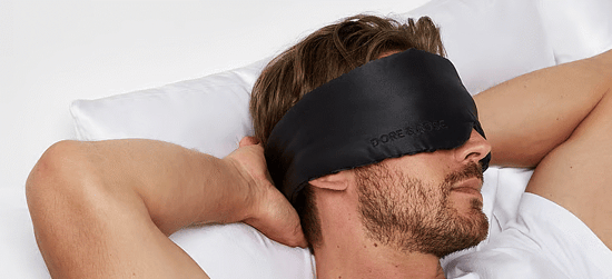 Mand ligger i en seng med armene bag hovedet og har en sort sovemaske over øjnene.