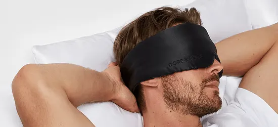 Mand ligger i en seng med armene bag hovedet og har en sort sovemaske over øjnene.