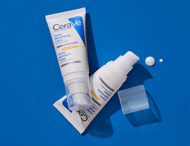 To CeraVe hudplejeprodukter ligger på en klar blå baggrund. En hvid tube med blå label ligger skråt oven på en flad emballage, og ved siden af ligger en pumpeflaske uden låg, hvor låget er placeret separat. Små dråber af creme ligger ved siden af produkterne. Kompositionen er dynamisk med tydelige skygger.