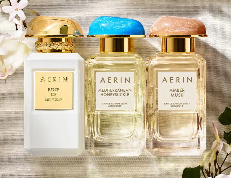 Her ses tre AERIN-parfumeflakoner: Rose de Grasse, Mediterranean Honeysuckle og Amber Musk. Flakonerne har guldfarvede detaljer og står side om side omgivet af blomster.
