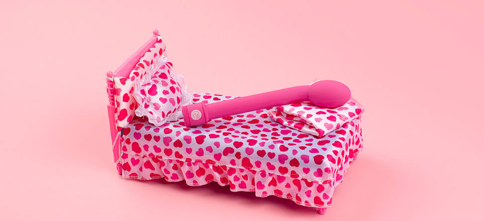 En legende og humoristisk opstilling på en ensfarvet, lyserød baggrund. I midten står en lille seng dækket af sengetøj med røde og pink hjerter på hvid bund, inklusive pude og tæppe. Oven på sengen ligger en stor, pink massage-stav (vibrator), som fylder det meste af sengen. Farverne er klare og kontrastfulde, og udtrykket er poppet og iøjnefaldende med fokus på produktet.