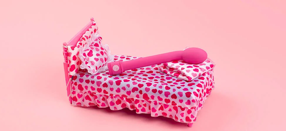 En legende og humoristisk opstilling på en ensfarvet, lyserød baggrund. I midten står en lille seng dækket af sengetøj med røde og pink hjerter på hvid bund, inklusive pude og tæppe. Oven på sengen ligger en stor, pink massage-stav (vibrator), som fylder det meste af sengen. Farverne er klare og kontrastfulde, og udtrykket er poppet og iøjnefaldende med fokus på produktet.