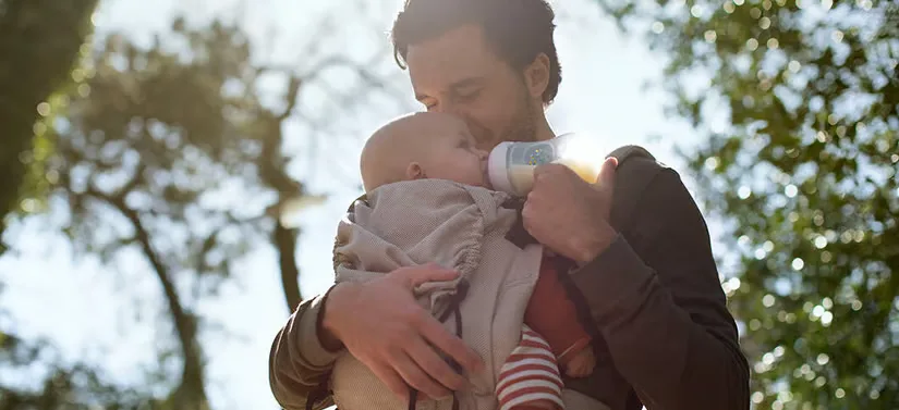 Her ses en far, som har sin lille baby i en holder på brystet. Faren holder en sutteflaske som babyen har i munden. De er ude i en skov.