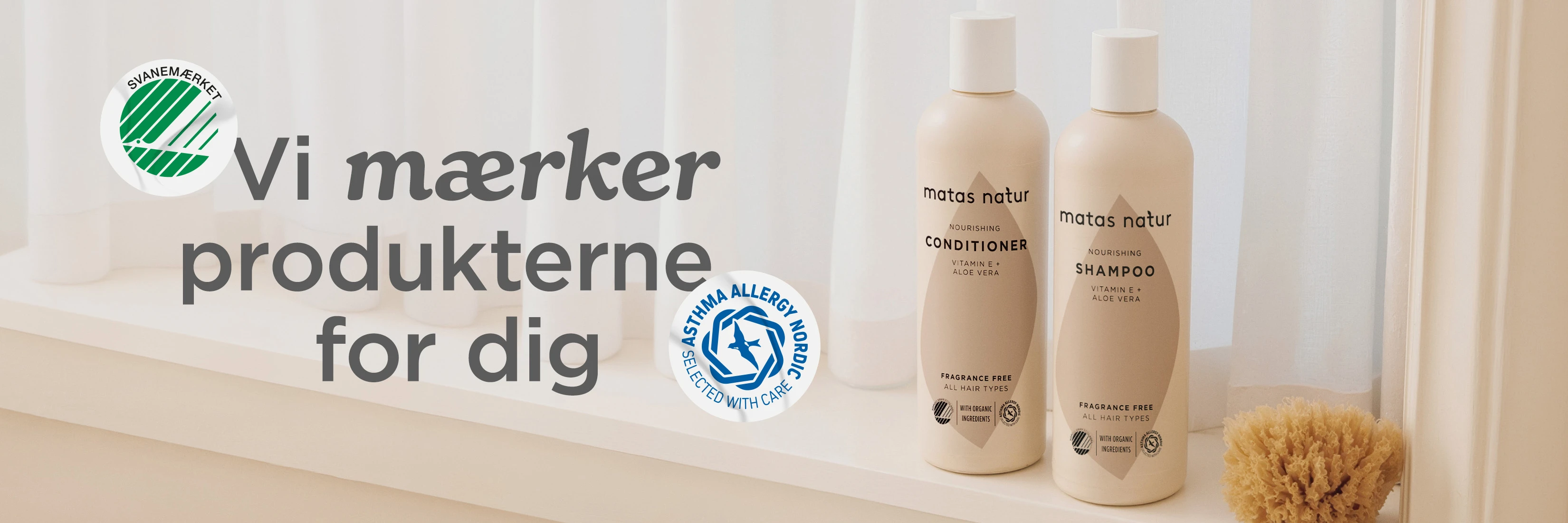 Her ses Matas Natur Shower Gel og Shampoo på en vindueskarm foran lyse gardiner. Ved siden af produkterne ligger en træbørste, et gråt håndklæde og en natursvamp. Billedet viser også Svanemærket, Asthma Allergy Nordic og A-kolben fra Forbrugerrådet Tænk.