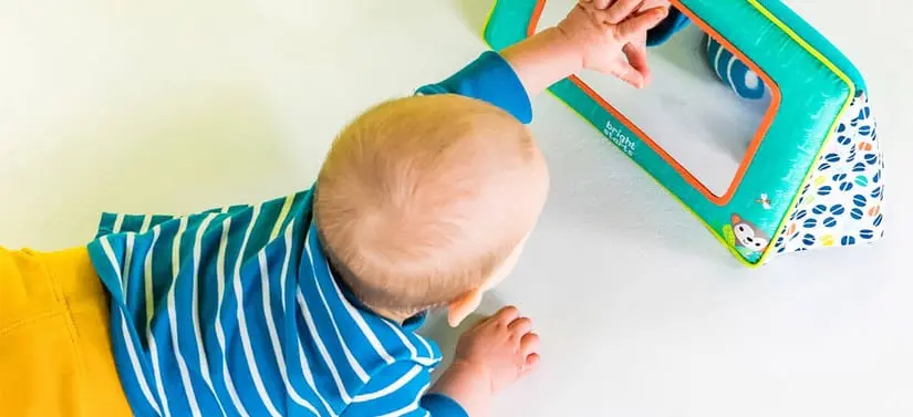 Baby ligger på maven på et lyst underlag og rækker hånden mod et farverigt stofspejl.