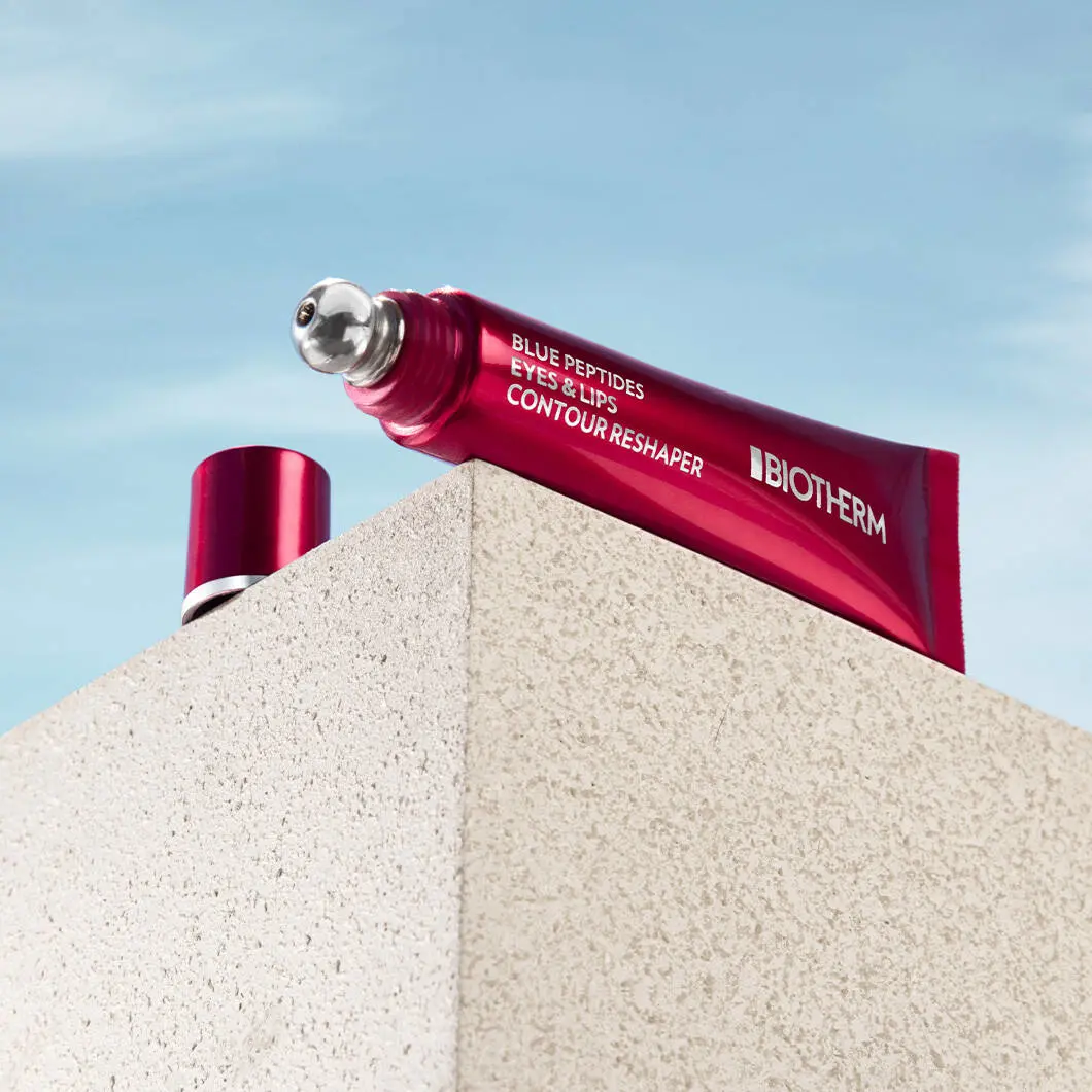 Et reklamebillede af et Biotherm hudplejeprodukt placeret på toppen af en lys stenblok mod en blå himmel med lette skyer. Produktet er en metallic rød tube med teksten “Blue Peptides Eyes & Lips Contour Reshaper” og Biotherms logo skrevet med hvide bogstaver. Tuben har en rund metalapplikator i spidsen, som er designet til at påføre produktet omkring øjne og læber. Ved siden af tuben ligger det røde låg. Billedet er taget nedefra, hvilket giver produktet et markant og eksklusivt udtryk. Det samlede look er moderne og luksuriøst med fokus på avanceret hudpleje og opstrammende pleje til ansigtets konturer.