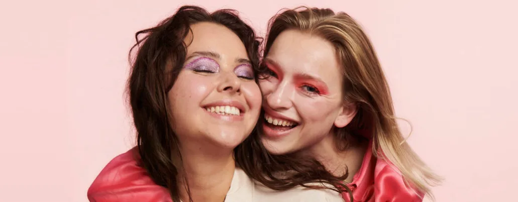 Her ses en lyserød baggrund med to glade kvinder. De har begge lyserød øjenmakeup på og smiler stort. 