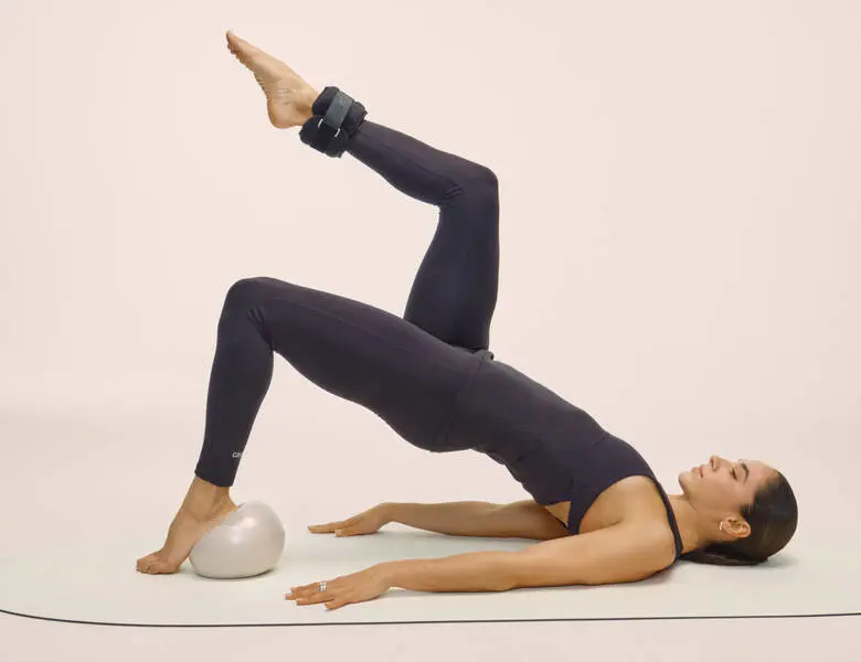 Her ses en kvinde i sort træningstøj, som ligger på ryggen ovenpå en lys yogamåtte. Hendes venstre ben er bukket og foden er oven på en hvid lille yogabold. Hun løfter numsen. Højre ben er løftet og bukket med en ankelvægt på