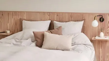 Lyst soveværelse med dobbeltseng, hvidt sengetøj, beige puder og sengegavl i træ.