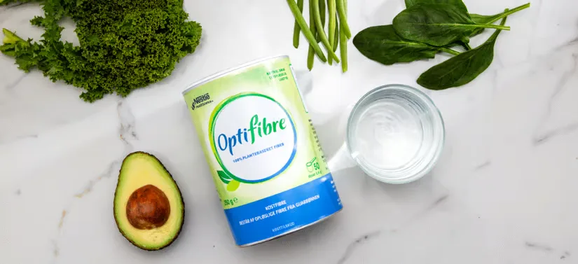 Her ses et OptiFibre produkt, som ligger på et hvidt bord. Rundt om er der forskellige grøntsager og et glad vand.