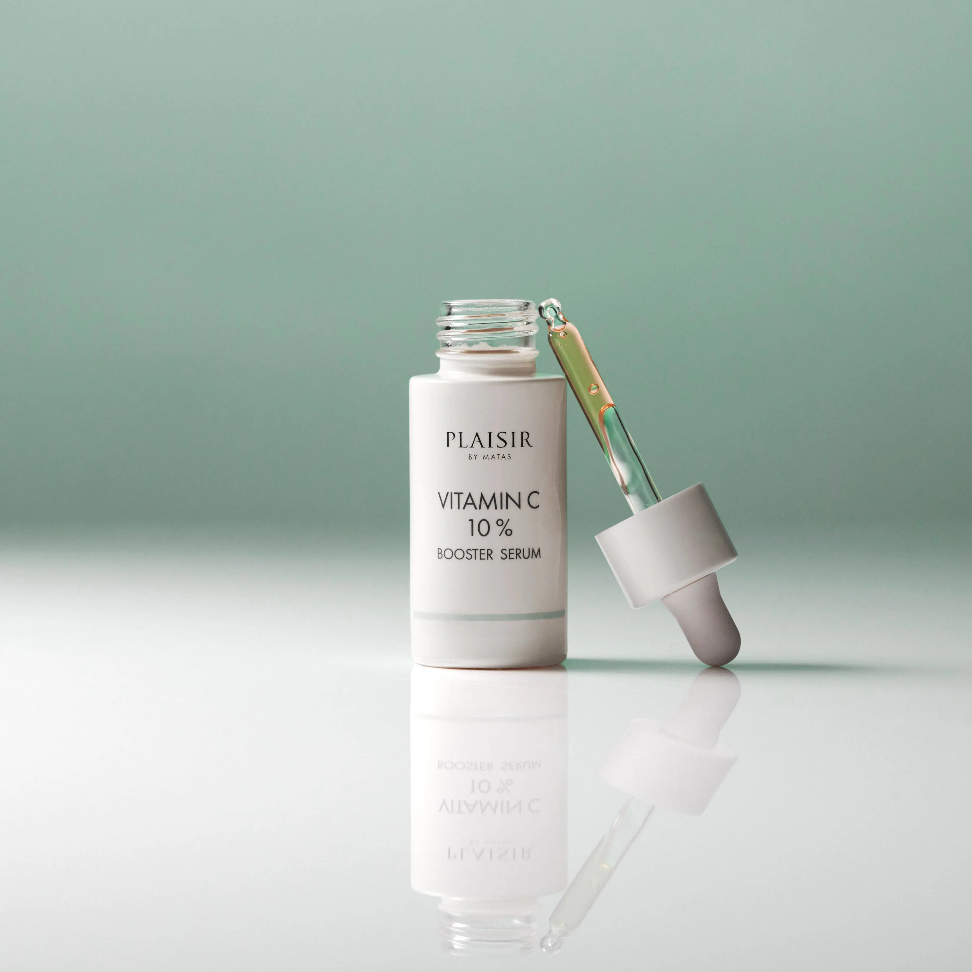 En flaske Plaisir Vitamin C 10% Booster Serum står åben på en blank overflade mod en grøn baggrund. Pipetten med gylden serum ligger ved siden af, og flasken spejler sig i underlaget.