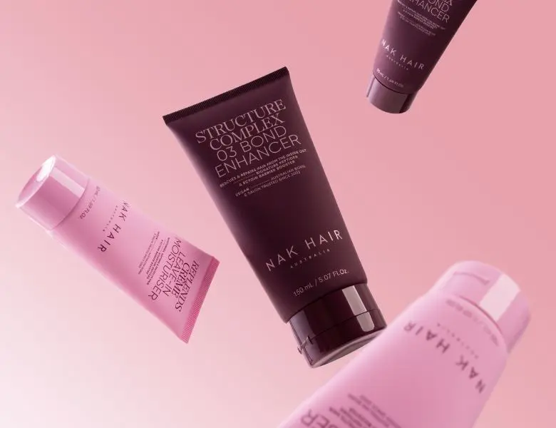 Her ses et billede med hårplejeprodukter fra NAK Hair placeret mod en lyserød baggrund. I midten ses produktet “Structure Complex No.3 Bond Enhancer”, omgivet af andre produkter fra NAK Hair-serien, herunder “Treatment Daily Detox Conditioner” og “Treatment Daily Detox Shampoo”.