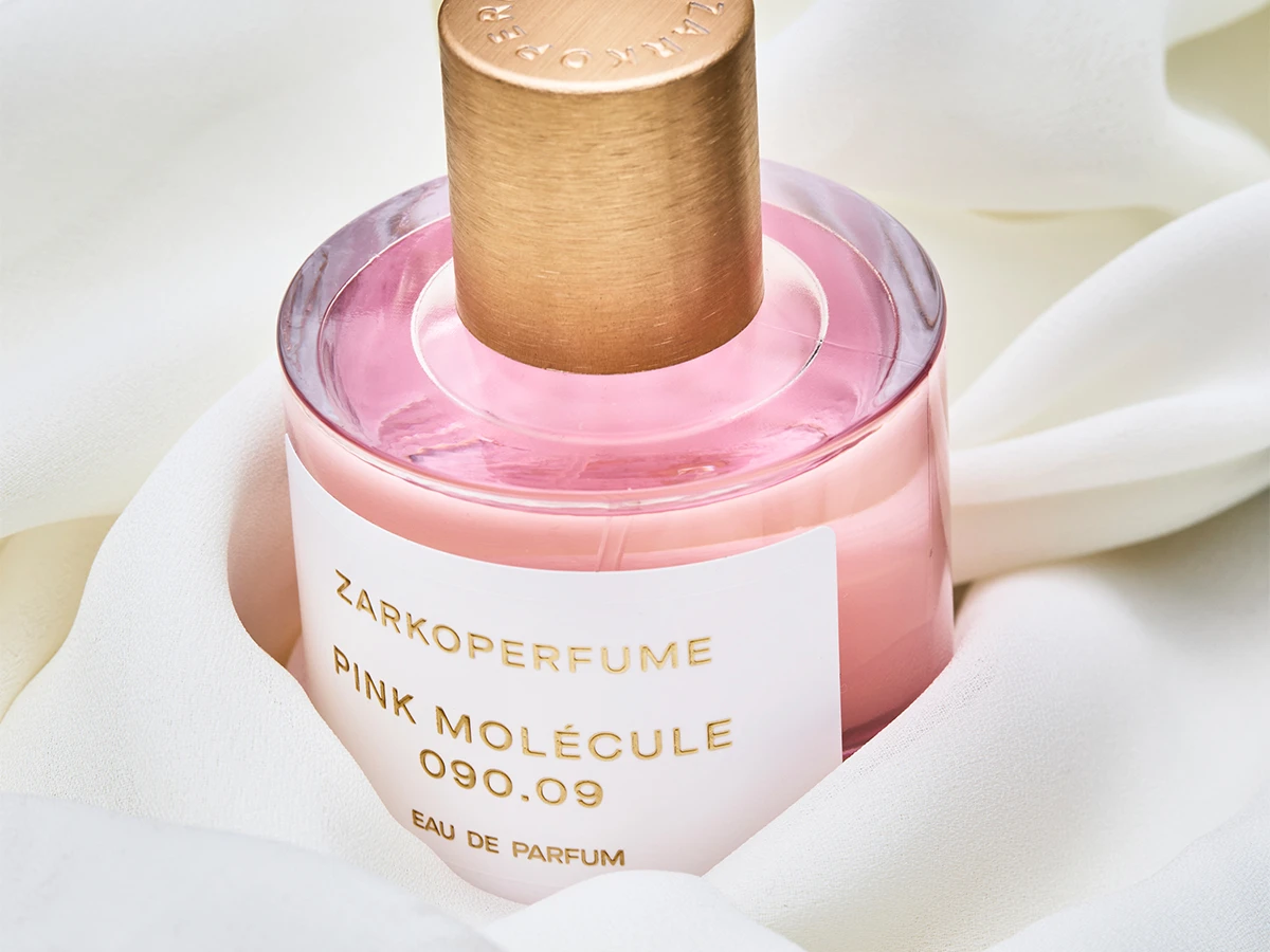 Her ses Zarkoperfume Pink Molécule 090.09 Eau de Parfum i en transparent parfumeflakon med rosa indhold, guldfarvet låg og hvid etiket. Flakonen er omgivet af lyst, bølgende tekstil.