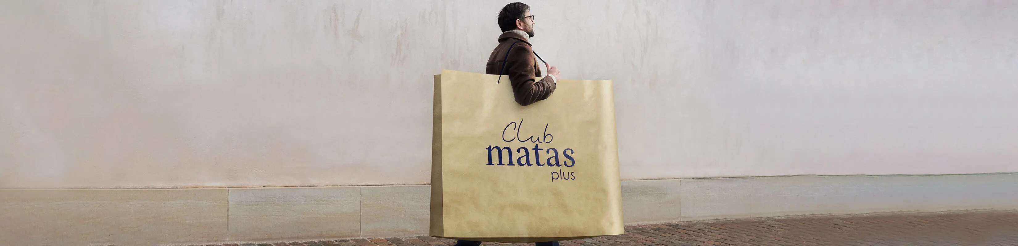 Her ses en mand skråt bagfra. Han går udenfor med en kæmpestor brun papirspose. På posen står der "Club Matas Plus" med blå skrift.
