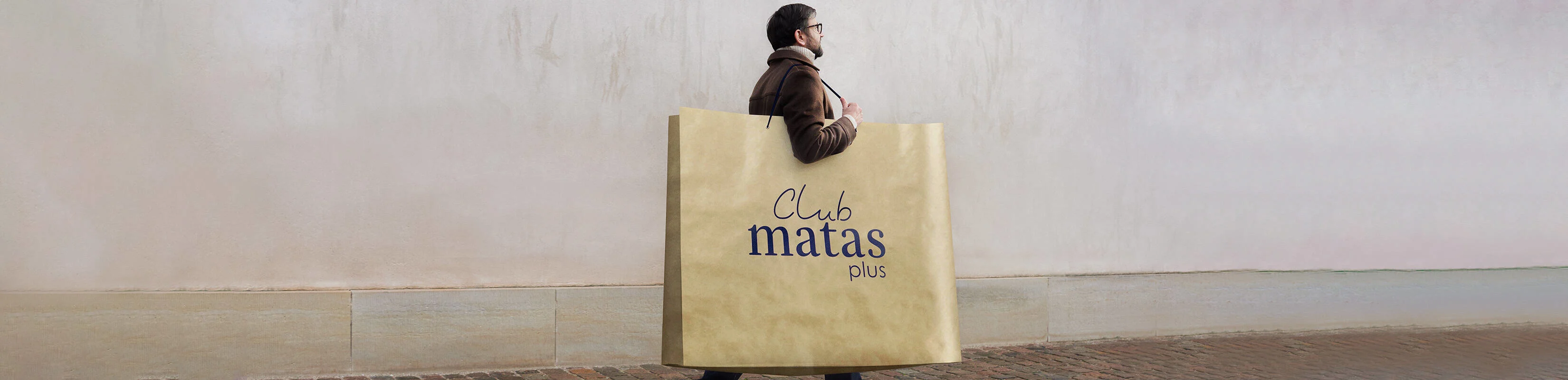Her ses en mand skråt bagfra. Han går udenfor med en kæmpestor brun papirspose. På posen står der "Club Matas Plus" med blå skrift.