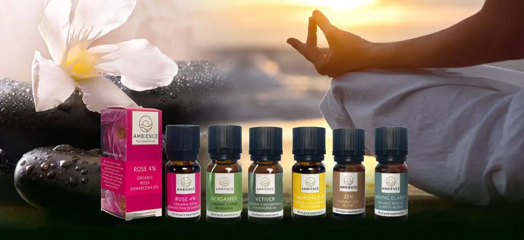 Her ses et udvalg af Ambience æteriske olier, blandt andet Rose, Bergamot, Vetiver, Chamomile, Zen og Mental Clarity. Produkterne står foran sten og en hvid blomst, mens en person mediterer i baggrunden ved solnedgang.
