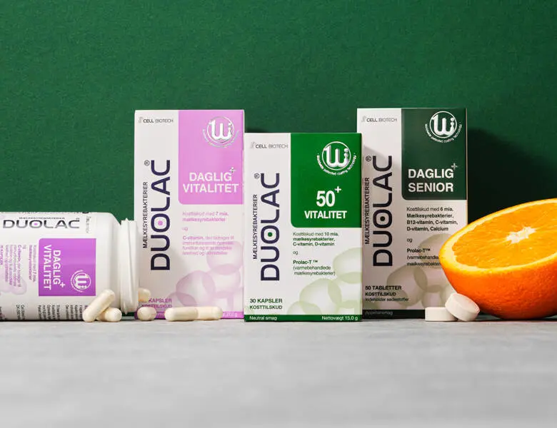 Duolac kosttilskud til daglig vitalitet, 50+ og senior står på grå overflade foran grøn baggrund sammen med kapsler og en halveret appelsin.