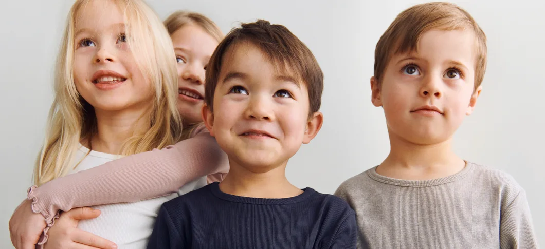 Fire børn står tæt sammen og smiler mod noget uden for billedet på en lys, neutral baggrund.