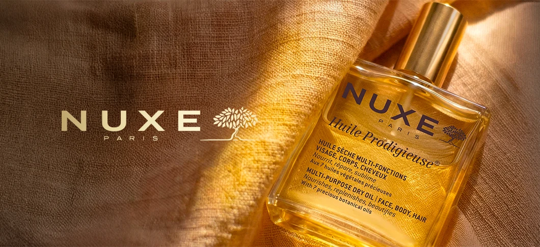 Her ses en NUXE Huile Prodigieuse olie i en gylden flaske på gyldent stof med teksten “NUXE Paris”.