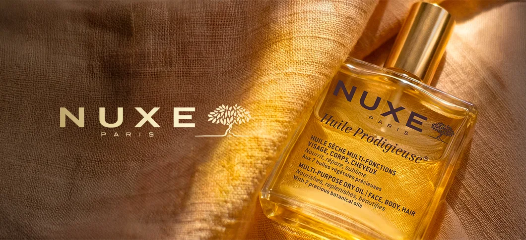 Her ses en NUXE Huile Prodigieuse olie i en gylden flaske på gyldent stof med teksten “NUXE Paris”.