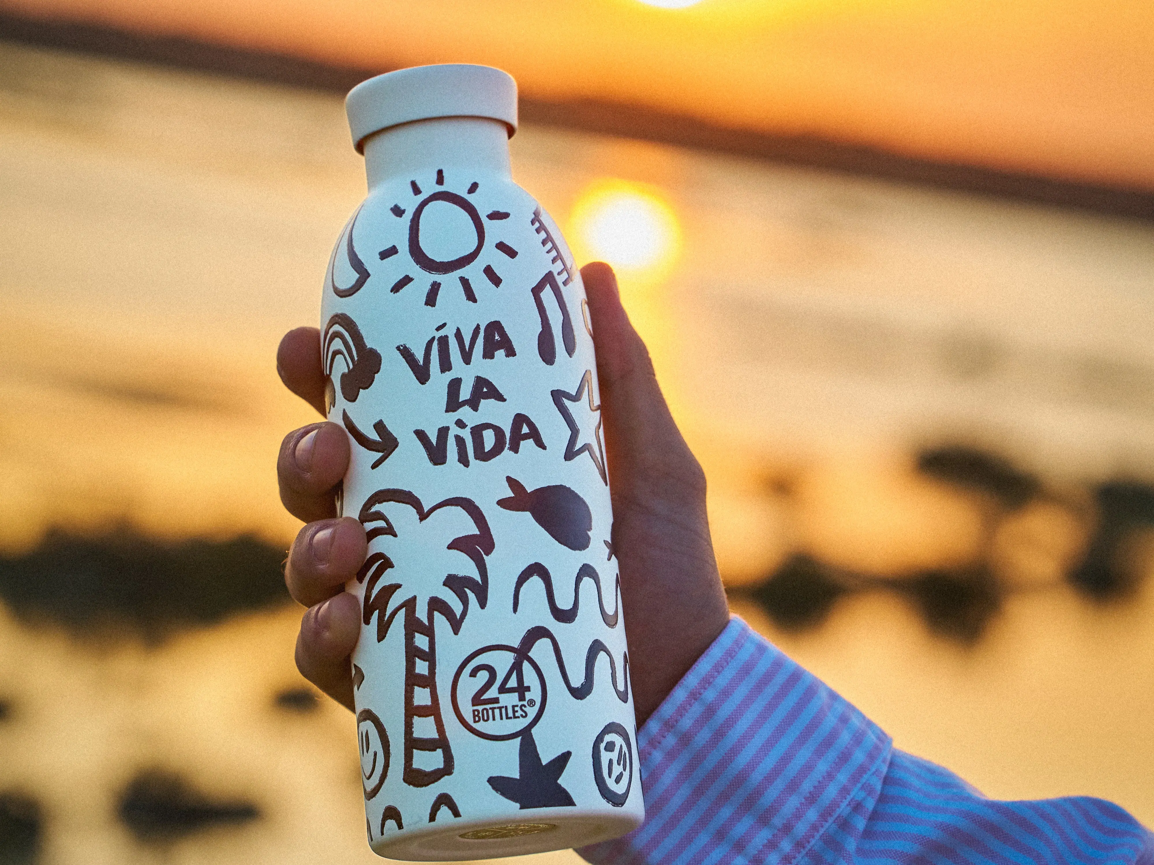 Hvid vandflaske med doodle-mønster, teksten 'VIVA LA VIDA' og '24 BOTTLES', holdes op mod en gylden solnedgang.