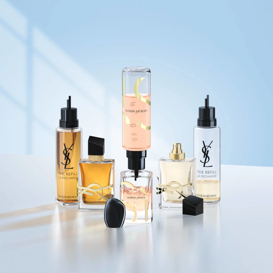 Fem luksusparfumer står samlet på en lys overflade foran en blød lyseblå baggrund. Produkterne fra Yves Saint Laurent og Giorgio Armani vises sammen med refillflasker monteret til genopfyldning. I midten fyldes en Giorgio Armani-parfume med lyserød væske, mens YSL Libre-flasker og refillbeholdere står på hver side. Flere låg ligger foran produkterne. Billedet har et elegant og moderne udtryk med fokus på refillbare parfumer og bæredygtige løsninger.
