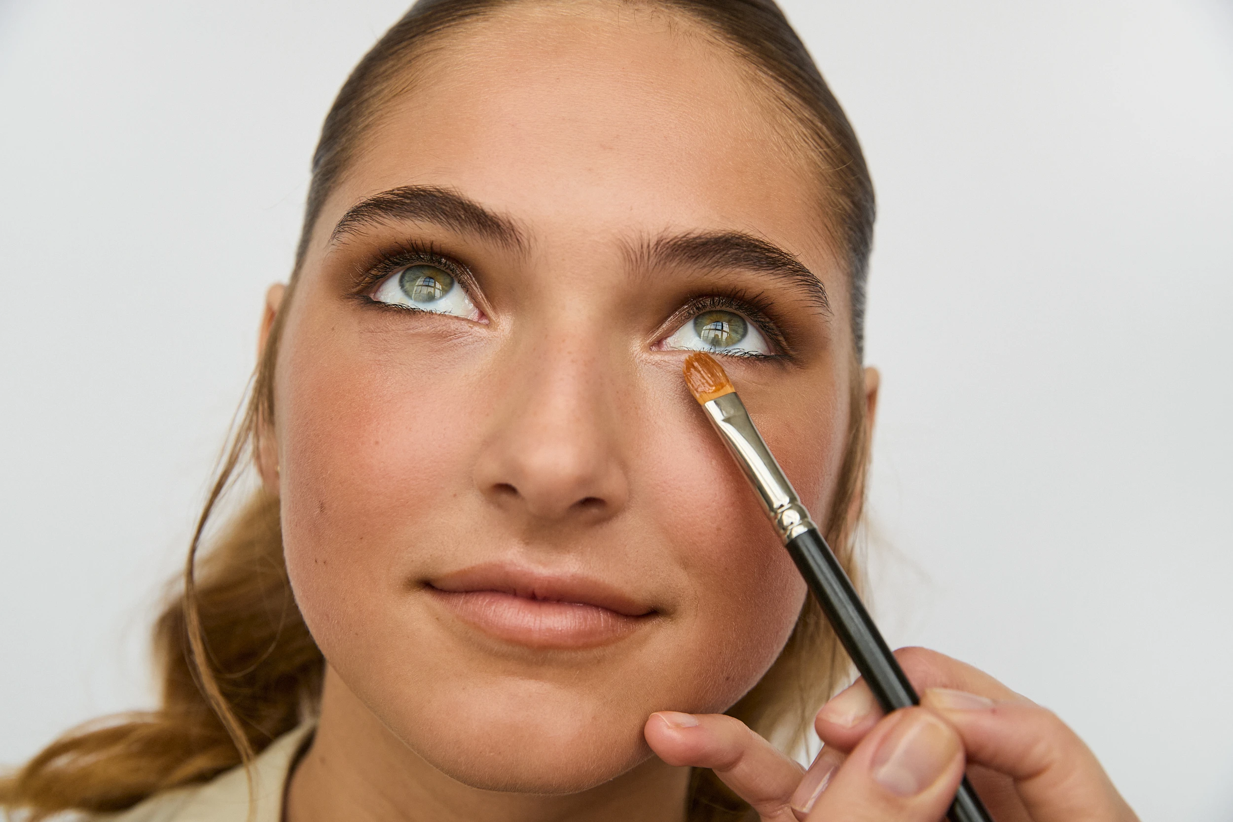 Her ses et nærbillede af en person, der får lagt makeup under øjet med en makeupbørste. Personen har opsat hår og makeup i brune og rosa nuancer og kigger opad mod en lys baggrund.