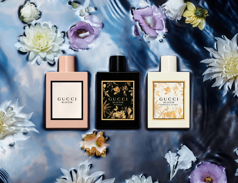 Her ses et billede med tre parfumeflakoner fra Gucci placeret side om side i vand med blomster omkring. På flakonerne står teksten “GUCCI BLOOM” samt “GUCCI BLOOM AMBROSIA DI FIORI”. Flakonerne er i lyserød, sort og lys farve med dekorative labels.
