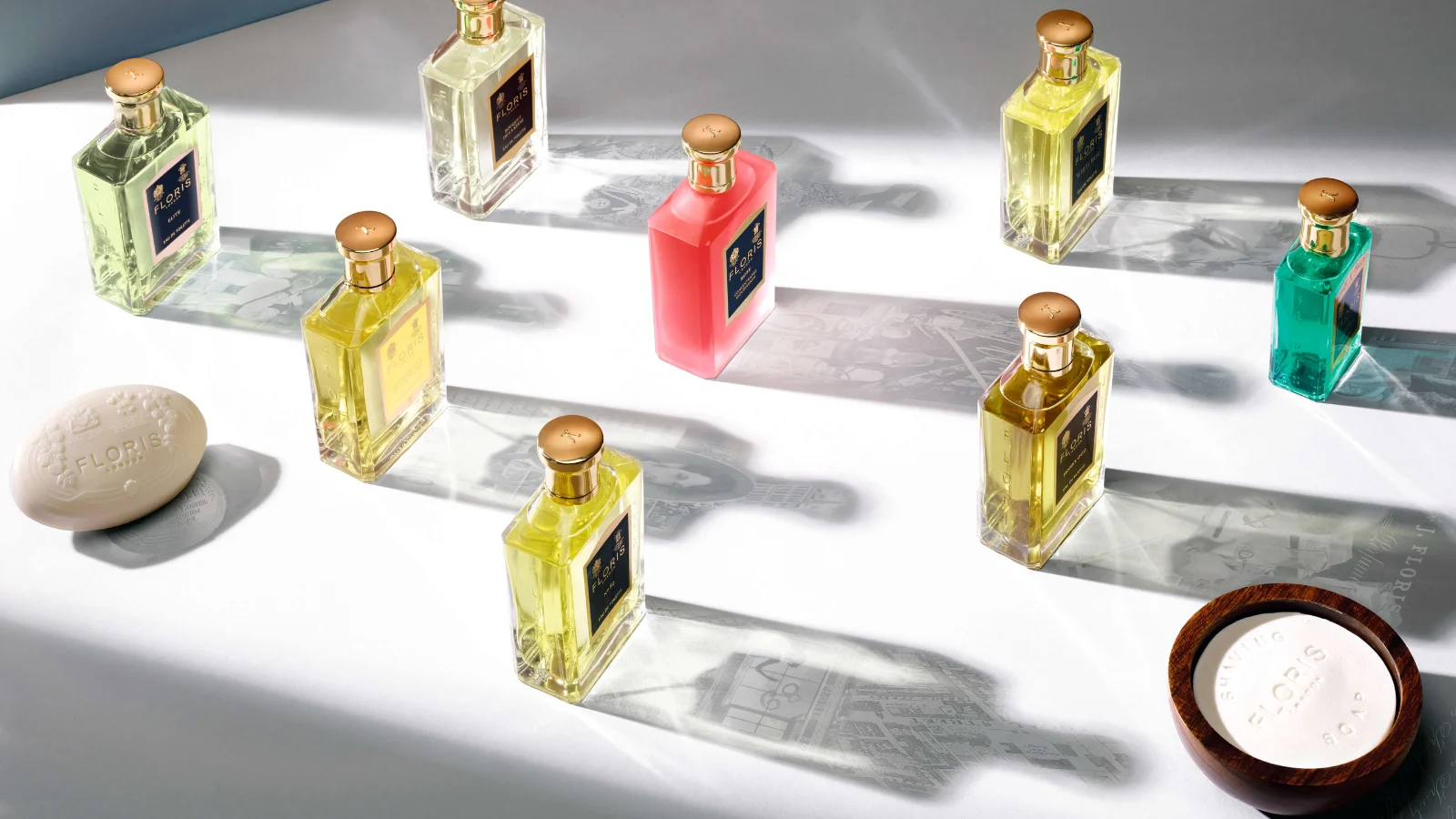 Flere Floris London parfumeflasker i forskellige farver står spredt i sollys på en hvid flade, omgivet af sæbe og dekorative detaljer.