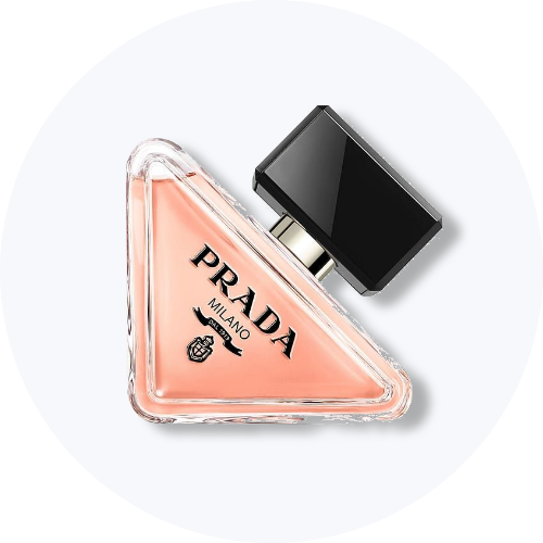 Her ses et billede af en parfumeflakon fra Prada placeret mod en lys baggrund. Flakonen har en trekantet form med lyserød nuance og sort låg, og på den står teksten “PRADA”.