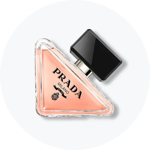 Her ses et billede af en parfumeflakon fra Prada placeret mod en lys baggrund. Flakonen har en trekantet form med lyserød nuance og sort låg, og på den står teksten “PRADA”.