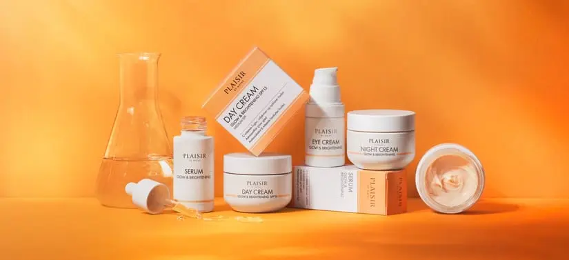 Reklamebillede med flere hudplejeprodukter arrangeret på en orange baggrund med varme lysnuancer. Produkterne består blandt andet af cremekrukker, serumflasker og æsker i hvide og beige farver med teksten “Flash”. Til venstre står en glasbeholder med gylden væske, og foran ligger en pipette med et par dråber olie. Produkterne er opstillet som et elegant stilleben med fokus på luksuriøs hudpleje og velvære.