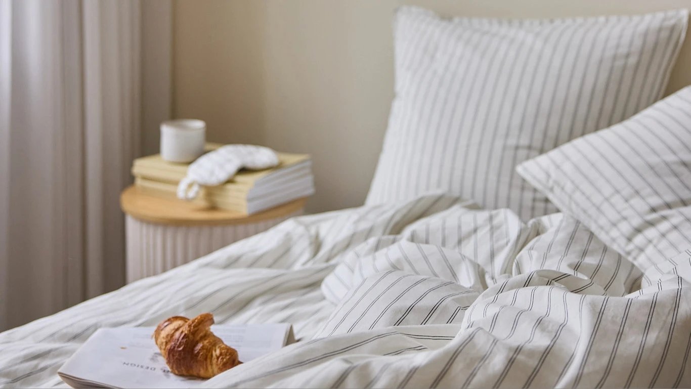 Her ses en seng med hvidt stribet sengetøj og puder. På sengen ligger en åben bog med en croissant, og på sidebordet står bøger, en kop og en sovemaske.