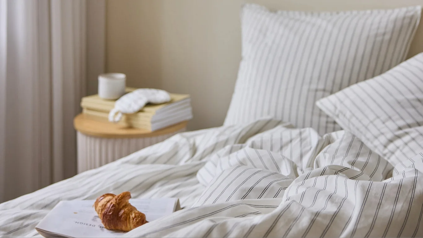 Her ses en seng med hvidt stribet sengetøj og puder. På sengen ligger en åben bog med en croissant, og på sidebordet står bøger, en kop og en sovemaske.