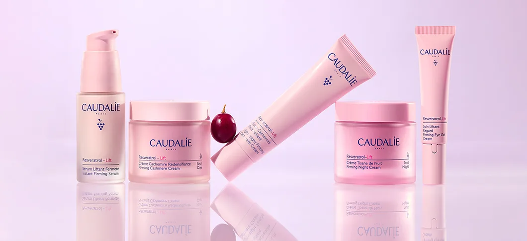 Her ses fem lyserøde Caudalie Resveratrol-Lift hudplejeprodukter og en vindrue på en lys lyserød baggrund med produkternes spejlinger i underlaget.