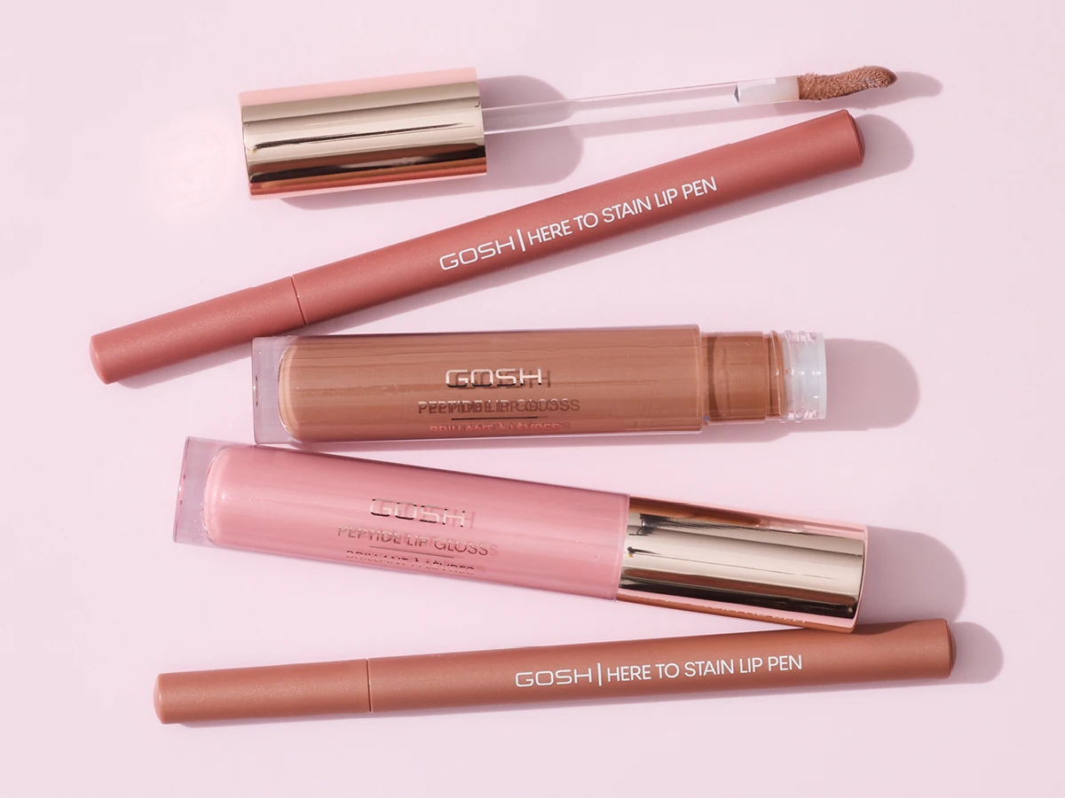 Her ses et udvalg af læbeprodukter fra GOSH Copenhagen mod en lyserød baggrund. Produkterne omfatter Here to Stain Lip Pen og Peptide Lip Gloss i forskellige rosa og nude nuancer.