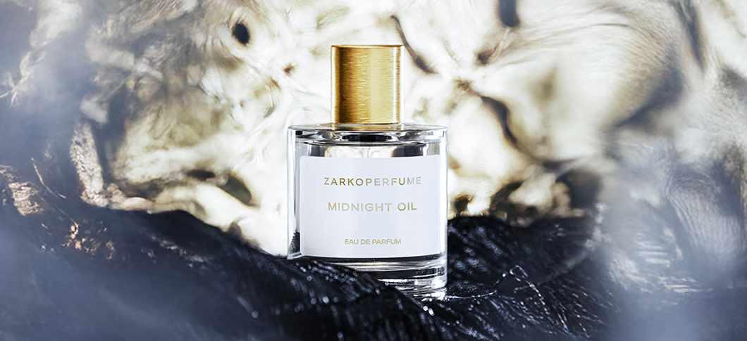 Her ses et billede af en parfumeflakon fra Zarkoperfume placeret centralt mod en mørk og abstrakt baggrund. På flakonen står teksten “ZARKOPERFUME MIDNIGHT OIL EAU DE PARFUM”. Flakonen er i klart glas med guldfarvet låg.